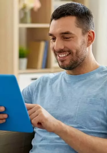 Homme souriant consultant une tablette numérique à domicile pour gérer ses traitements médicaux.