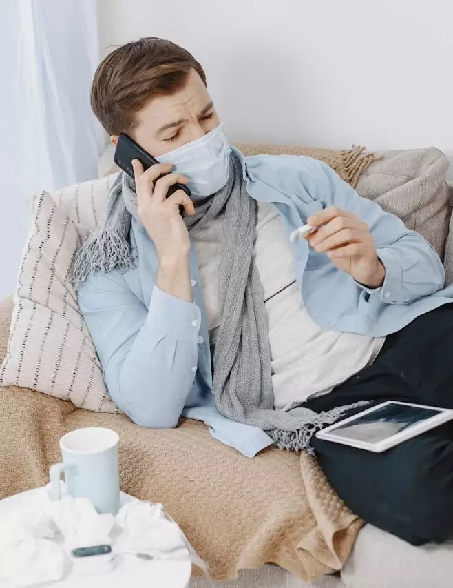 Homme malade portant un masque, prenant sa température et téléphonant depuis son canapé à domicile.