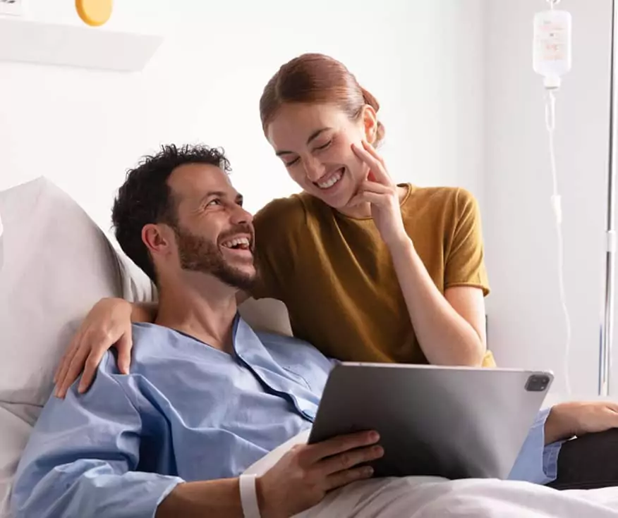 Homme souriant en milieu hospitalier, accompagné d’une femme, consultant ensemble une tablette numérique.